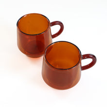 Load image into Gallery viewer, Two amber-brown translucent glass mugs with handles, photographed on a clean white background.
